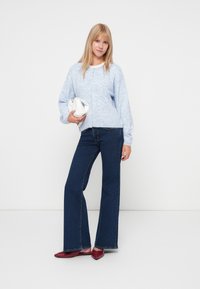 Lyseblå strikket cardigan med knapper, parret med mørkeblå vidbenede jeans og røde flade sko. Personen holder en hvid taske.