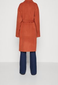 Persona che indossa un cappotto di lana color ruggine con cintura e jeans a gamba larga blu scuro, vista da dietro su uno sfondo bianco uniforme.