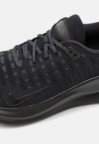 Sort sportsko med en strikket overdel, struktureret mønster og glat gummisål. Har sorte snørebånd og et sort Nike swoosh-logo.