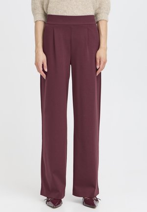 Femeie purtând pantaloni plum cu talie înaltă și croială largă, asortați cu un pulover bej tricotat și pantofi plum cu vârf ascuțit, stând în fața unui fundal alb simplu.