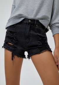 Shorts de mezclilla negra desgastada con rasgaduras y dobladillos deshilachados, combinados con una blusa de manga larga gris, mostrados desde la mitad del muslo hasta la cintura.