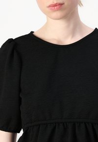 Top in tessuto nero a trama con maniche corte a palloncino e scollatura rotonda, caratterizzato da una vita arricciata e un delicato dettaglio a volant all'orlo.