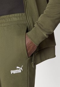 Veste de sport vert olive avec une poche zippée et un pantalon de jogging assorti. Logo PUMA en blanc sur la jambe. Texture de tissu douce.
