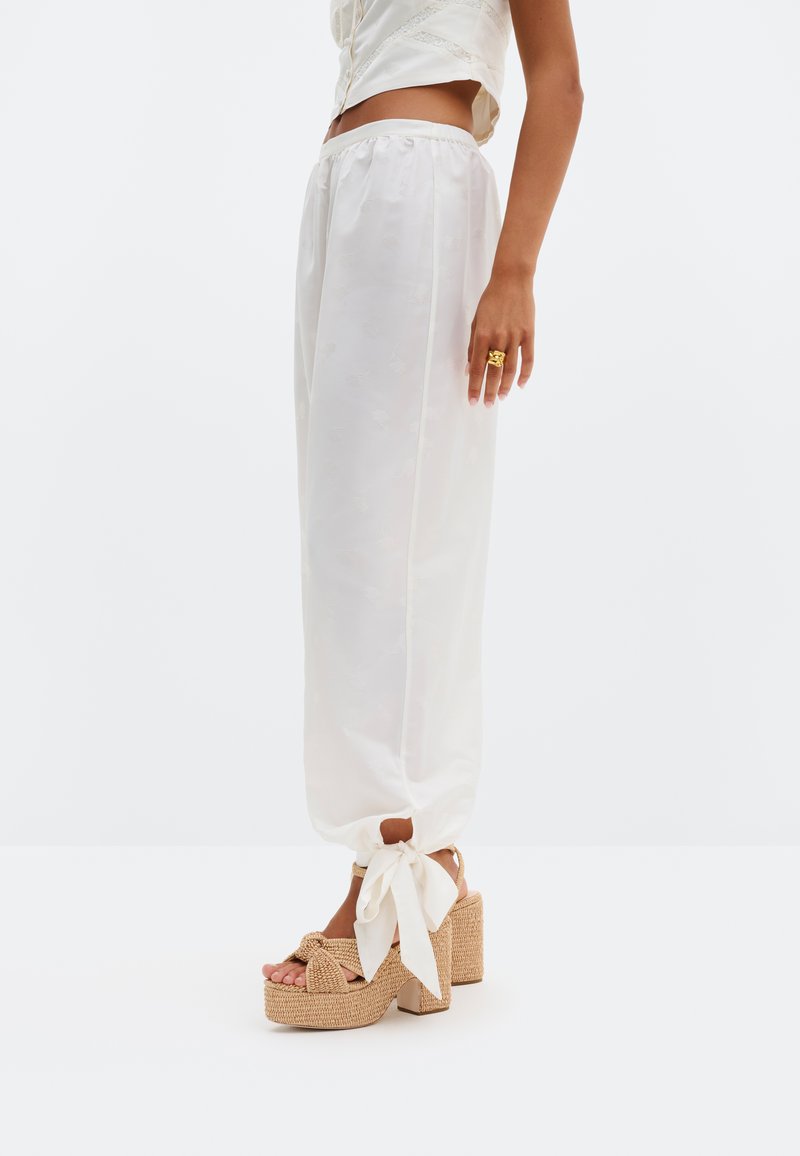 Γυναίκα που φοράει λευκό σατέν παντελόνι με κορδόνια στον αστράγαλο και μπεζ υφασμάτινα πλατφόρμα τακούνια, στέκεται μπροστά σε λευκό φόντο.
