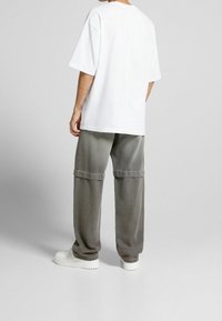 Person klädd i en överdimensionerad vit T-shirt, lösa grå cargo-byxor med sömdetalj ovanför knäna, samt vita plattformssneakers, stående på en grått golv.