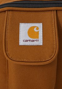 Carhartti kaubamärgiga kangas, pruun kanvas koos ristkülikukujulise plaadi ning kollase logo ja tekstiga, kõva tõmblukk üleval. Tekstuuriga pind.