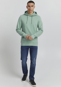 Lichtgroene hoodie met trekkoorden, kangaroozak, ribgebreide manchetten, gedragen met donkerblauwe jeans en zwarte sneakers. Zachte stoftextuur.