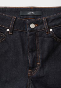 Dark blue denim jeans with orange stitching, metal button and rivets, and a label reading "someday." sewn inside the waistband.