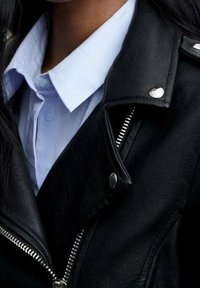 Persona che indossa una camicia azzurro chiaro con colletto sotto una giacca di pelle nera con zip in argento visibili e bottoni a pressione.