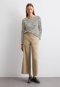 Gestreiftes Langarmhemd in Creme und Schwarz; beigen, weiten Hosen mit Fronttaschen; schwarze Slippers; strukturierte Oberfläche.