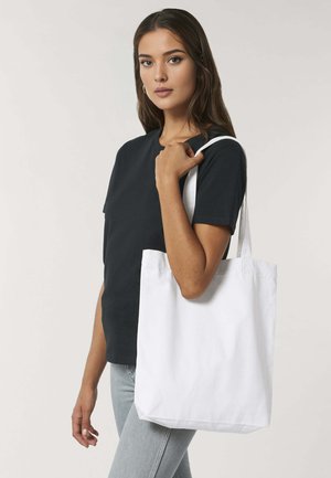Witte katoenen tote bag met twee schouderbanden, rechthoekige vorm en een eenvoudige hoeveelheid, vastgehouden door een persoon die een zwart T-shirt draagt.