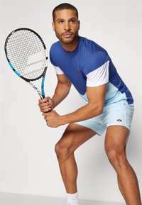 Blaues und weißes Sportshirt mit hellblauen Shorts, hält einen Tennisschläger, mit einem Logo auf dem Shirt und den Shorts.