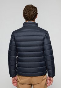 Chaqueta acolchada de color azul marino con cuello alzado, diseño acolchado horizontal y puños elásticos. Textura suave, aislamiento ligero visible.