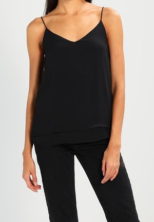 Kvinde iført en ærmeløs sort V-hals camisole med tynde stropper og sorte bukser foran en ensfarvet lys baggrund.