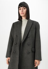 Femme aux cheveux noirs raides portant un manteau gris ardoise croisé et un col roulé à rayures claires, se tenant devant un fond blanc.