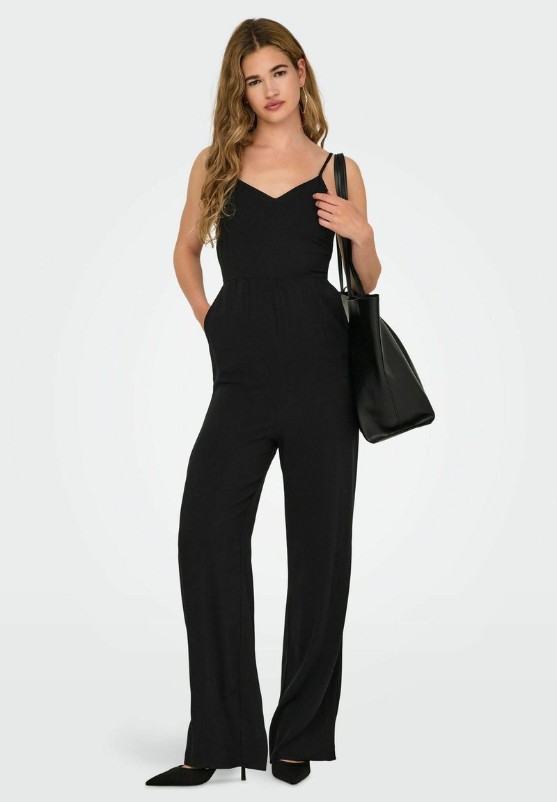 Zwarte jumpsuit met een getailleerde corsage, v-hals, ruime broeken en zijzakken. Vergezeld van een zwarte handtas en zwarte puntige hakken.
