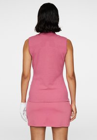 Ärmlös rosa golftröja med hög krage, texturerad baksida, figurnära design och tillsammans med en matchande rosa kjol. Kompletteras med en vit handske.