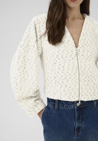 Cremefarvet lynlåstrøje med strukturerede skuldre, tekstureret stof og en dyb V-hals, kombineret med højtaljede blå denimjeans.