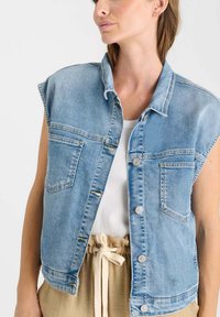 Leren mouwloos vest in lichtblauw met twee voorzakken, een knoopsluiting aan de voorkant en een kraag. Gedragen over een witte top.