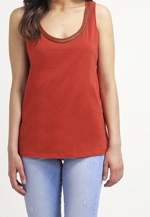 Kobieta ubrana w bezrękawnik w kolorze rdzy z ozdobnym wykończeniem na dekolcie oraz jasnoniebieskie jeansy z przetarciami, stojąca na tle jednolitego białego tła.