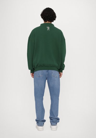 Sweatshirt vert surdimensionné avec un col haut et un petit détail brodé au dos, associé à un jean droit bleu clair.