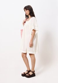 Weißes Leinenkleid mit strukturierten Streifen, kurzen Ärmeln und Spitzenakzenten. Kombiniert mit dunkelbraunen Plateausandalen und einer rosa geflochtenen Tasche.