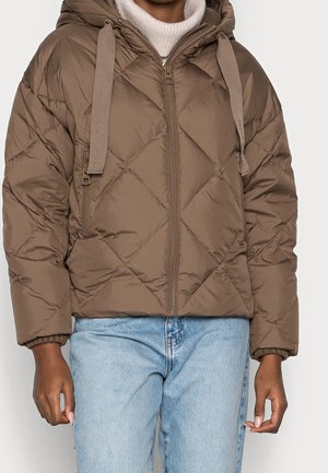 Bruine doorgestikte jas met ritssluiting, capuchon en brede trekkoorden, gedragen over een witte coltrui, gecombineerd met lichtblauwe spijkerbroek.