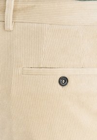 Beżowe spodnie sztruksowe z pionowymi prążkami, z pojedynczą kieszenią i czarnym guzikiem na linii talii.