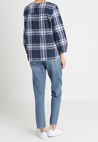 Camicia a quadri blu navy con linee bianche e grigie, tessuto leggero, vestibilità ampia, scollatura rotonda e maniche lunghe a sbuffo, abbinata a jeans in denim chiaro.