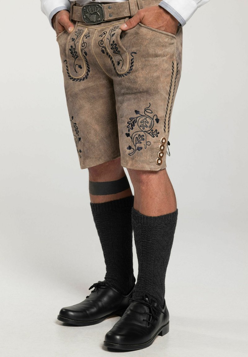 Braune Lederhosen mit kunstvoller blauer Blumenstickerei, ausgestattet mit Seitentaschen und Knöpfen, kombiniert mit schwarzen knielangen Socken und Schuhen.