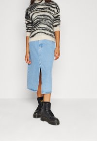 Pull en tricot avec un motif tourbillonnant noir et beige, associé à une jupe midi en denim bleu avec une fente à l'avant et des bottes noires à lacets.