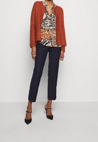 Cardigan di color ruggine, texturizzato, indossato su una blusa decorata con motivi marroni, neri e bianchi; abbinato a pantaloni blu navy e tacchi neri.