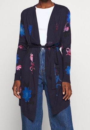 Personne portant un cardigan bleu marine foncé avec des imprimés floraux bleus et roses, ceinturé à la taille, sur un top blanc et un jean bleu.