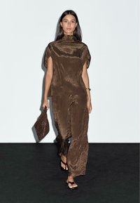Top a pieghe marrone e pantaloni palazzo in un tessuto fluido, simile alla satin. Abbinati a una borsa a tracolla testurizzata e sandali con cinturino. Design minimalista.
