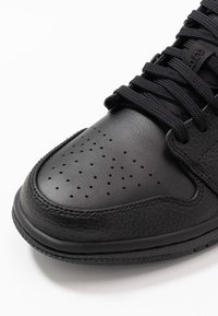 Gros plan sur le bout d'une basket en cuir noir avec des perforations, des coutures texturées et des lacets noirs sur fond blanc.