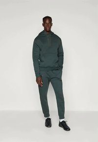 Vīrietis stāv tumši zaļā kapucē un atbilstošos biksēs ar melnām sporta kedām uz vienkārša gaiša fona.