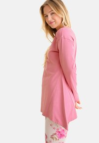 Tunique rose à manches longues avec un ourlet arrondi, associée à des leggings à motif floral. Le tissu semble lisse et léger.