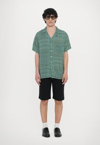 Camisa de manga curta, com padrão verde e bolso, combinada com calções pretos. O modelo usa óculos de sol escuros e mocassins pretos.