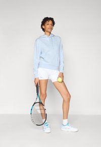 Felpa blu chiaro, pantaloni corti bianchi, tiene una racchetta da tennis e una pallina. Presenta una tasca a marsupio e un cappuccio con coulisse. Scarpe da ginnastica con accenti blu.