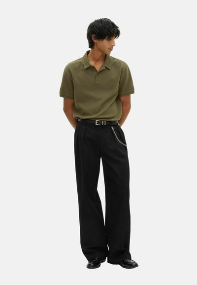 Giovane uomo che indossa una polo a maniche corte verde oliva, pantaloni neri a gamba larga con accessorio a catena, scarpe nere, in piedi con le mani dietro la schiena.