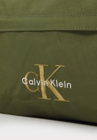 Vihreä kangaskassi, jossa on brodeerattu "Calvin Klein" -teksti valkoisena ja kultaisena, tasaisella pinnalla ja siistillä, modernilla muotoilulla.