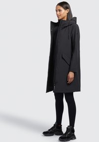 Parka nera impermeabile con cappuccio, chiusura con zip, tasche frontali e vestibilità rilassata, abbinata a leggings neri e sneakers chunky.