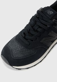 Czarne sportowe buty z teksturowanym syntetycznym skórzanym cholewką, sekcjami z siateczki i ciasno tkanymi sznurowadłami; cechują się wyraźnym logo "N" i białą podeszwą.