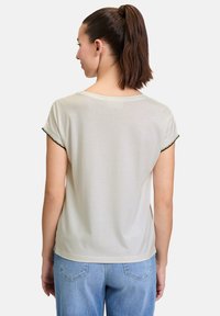 Lyse beige t-shirt med korte ærmer, med rund halsudskæring og dekorativ sort syning langs ærmeenderne. Blødt bomuldsstof.