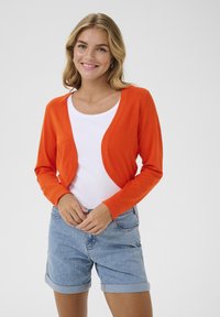Cardigan lavorato a maglia arancione con maniche lunghe e design aperto; abbinato a una camicia bianca e a pantaloni shorts in denim blu, che mettono in risalto uno stile casual.