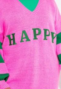 Roze gebreide trui met een V-hals, voorzien van groene strepen op de mouwen en het woord "HAPPY" in glinsterende groene letters over de voorkant.