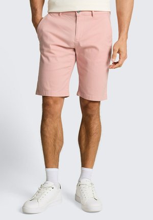 Man draagt lichtroze knielange shorts, witte sokken en witte sneakers tegen een egale lichte achtergrond.