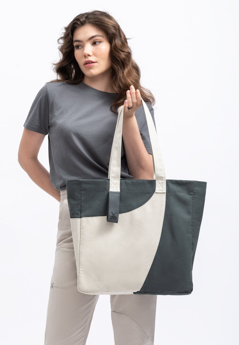 Canvas draagtas in crème en donkergroen met een gebogen ontwerp. Voorzien van stevige handvatten en een label met een logo. Model houdt de tas casual vast.