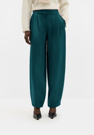 Femme portant un pantalon taille haute, large et plissé, de couleur bleu canard, associé à des chaussures noires à bout pointu et un pull crème duveteux.