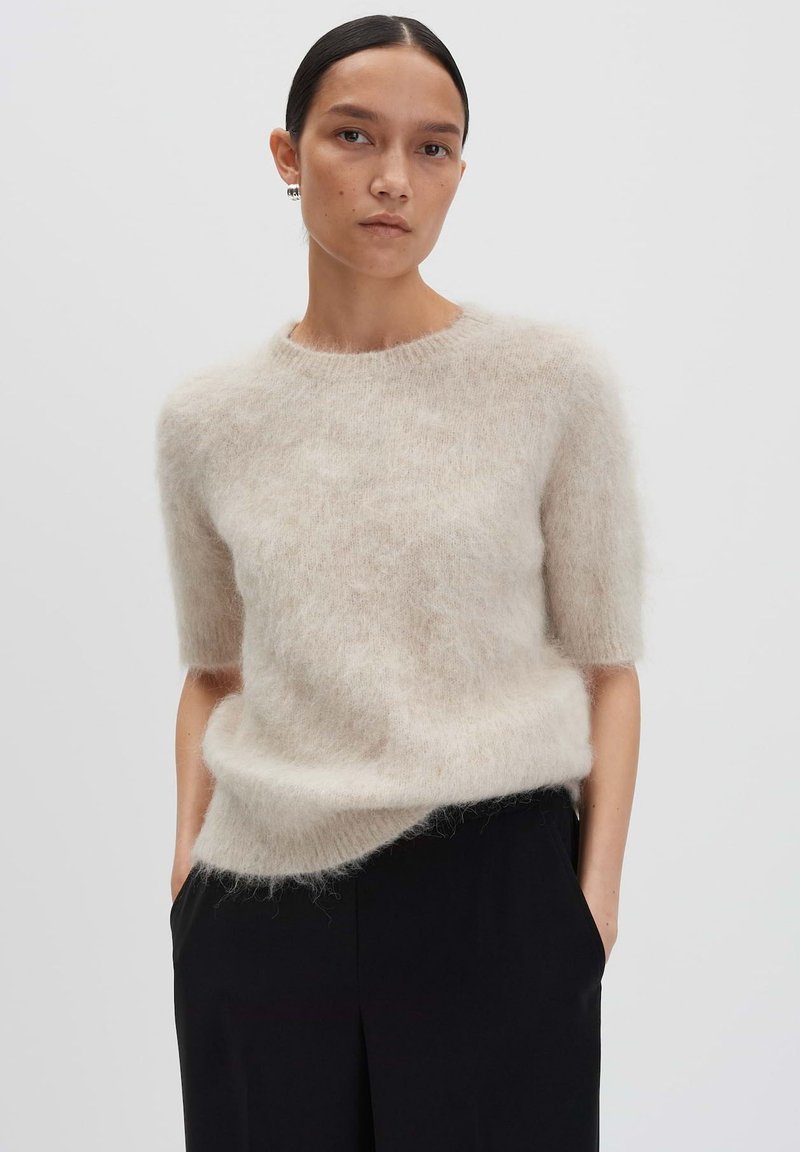 Pull léger beige à manches courtes en matière moelleuse avec un col rond et une coupe décontractée, associé à un pantalon noir.